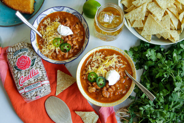 Bowls of chili topped with cheese, jalapeños, and sour cream sit on a table. Surrounding are tortilla chips, fresh cilantro, a lime, a drink, and a bag of pinto beans.