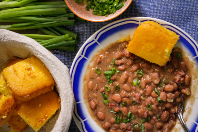 A close-up of a plate with savory beans garnished with chopped green onions, accompanied by a piece of golden cornbread. Nearby, more cornbread in a basket and fresh green onions.