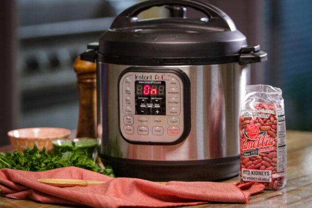 Instant Pot with digital display set to "On" sits on a counter. Nearby are a bag of red kidney beans, fresh herbs, and a wooden spoon on a red cloth.