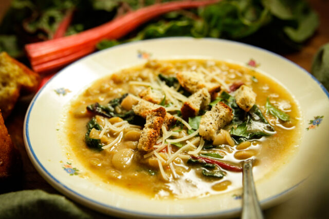 A warm bowl of soup with chickpeas, leafy greens, and shredded cheese, garnished with croutons. The dish is served in a floral-trimmed bowl.