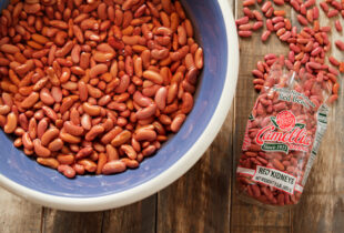 A bowl filled with soaking red kidney beans next to a Camellia brand bag of dry beans on a rustic wooden table, conveying a homely kitchen feel.