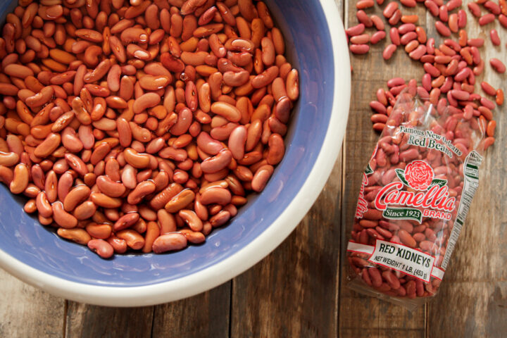 A bowl filled with soaking red kidney beans next to a Camellia brand bag of dry beans on a rustic wooden table, conveying a homely kitchen feel.