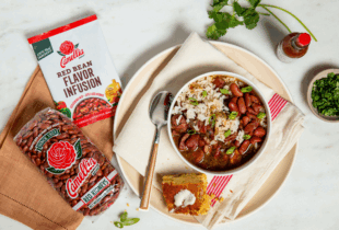 Bowl of red beans and rice with green onions on a napkin-lined plate, next to cornbread with butter, Camellia beans and flavor mix, hot sauce, and herbs.