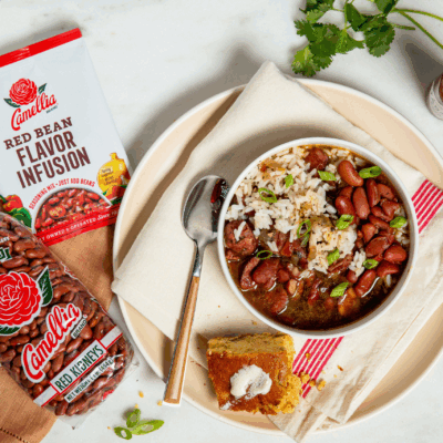 Bowl of red beans and rice with green onions on a napkin-lined plate, next to cornbread with butter, Camellia beans and flavor mix, hot sauce, and herbs.