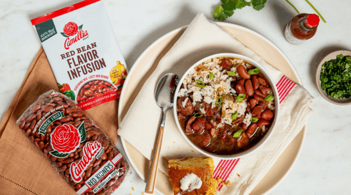 Bowl of red beans and rice with green onions on a napkin-lined plate, next to cornbread with butter, Camellia beans and flavor mix, hot sauce, and herbs.