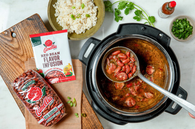 A pot of red beans with sausage in sauce, being ladled. Nearby are a bag of dry beans, a seasoning packet, cooked rice in a bowl, and fresh cilantro.