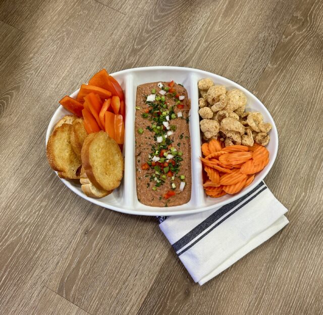 Oval platter with sliced baguette, carrot sticks, diced red peppers, and a pâté topped with herbs and onions. Casual dining setup on wood surface.