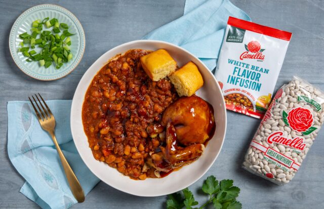 A bowl of saucy baked beans with chicken and cornbread on a blue napkin. Nearby are chopped green onions, a fork, and Camellia white beans packaging.