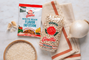 Package of Camellia white beans and seasoning mix on a marble surface, surrounded by garlic cloves, an onion, rice, and a beige kitchen towel.