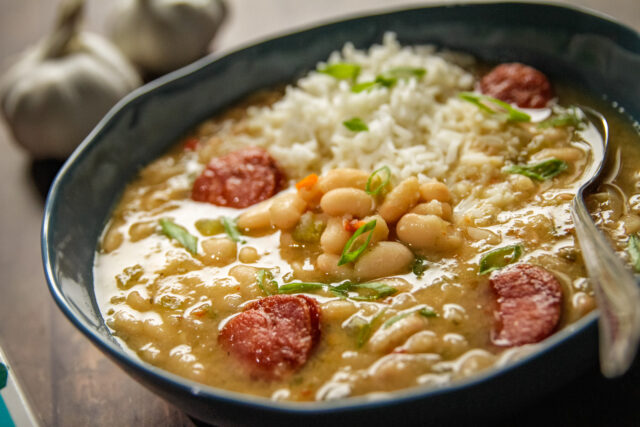 A hearty bowl of bean soup with sliced sausage, white rice, and green onions, served in a blue dish. Two garlic bulbs are blurred in the background.