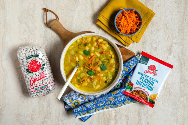 A vibrant bowl of white bean soup garnished with cilantro and carrots sits on a wooden board. Nearby are a packet of beans and seasoning mix. Warm and inviting.