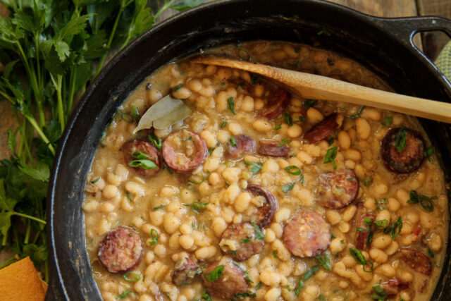 Hearty bean stew with sliced sausage in a black pot, garnished with chopped herbs. A wooden spoon rests on top, evoking warmth and comfort.