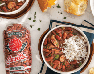 A bowl of red beans and rice is garnished with green onions, accompanied by cornbread with butter. Nearby are Camellia red bean packaging and cilantro.