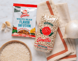 A bag of Camellia Great Northern beans and a white bean flavor infusion packet are on a table with garlic, a white onion, rice, and a cloth napkin.