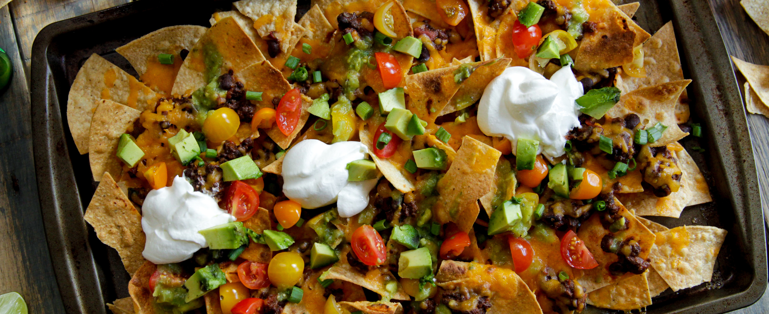 A tray of vibrant nachos topped with melted cheese, black beans, diced avocado, cherry tomatoes, scallions, and dollops of sour cream, evoking a festive, appetizing atmosphere.