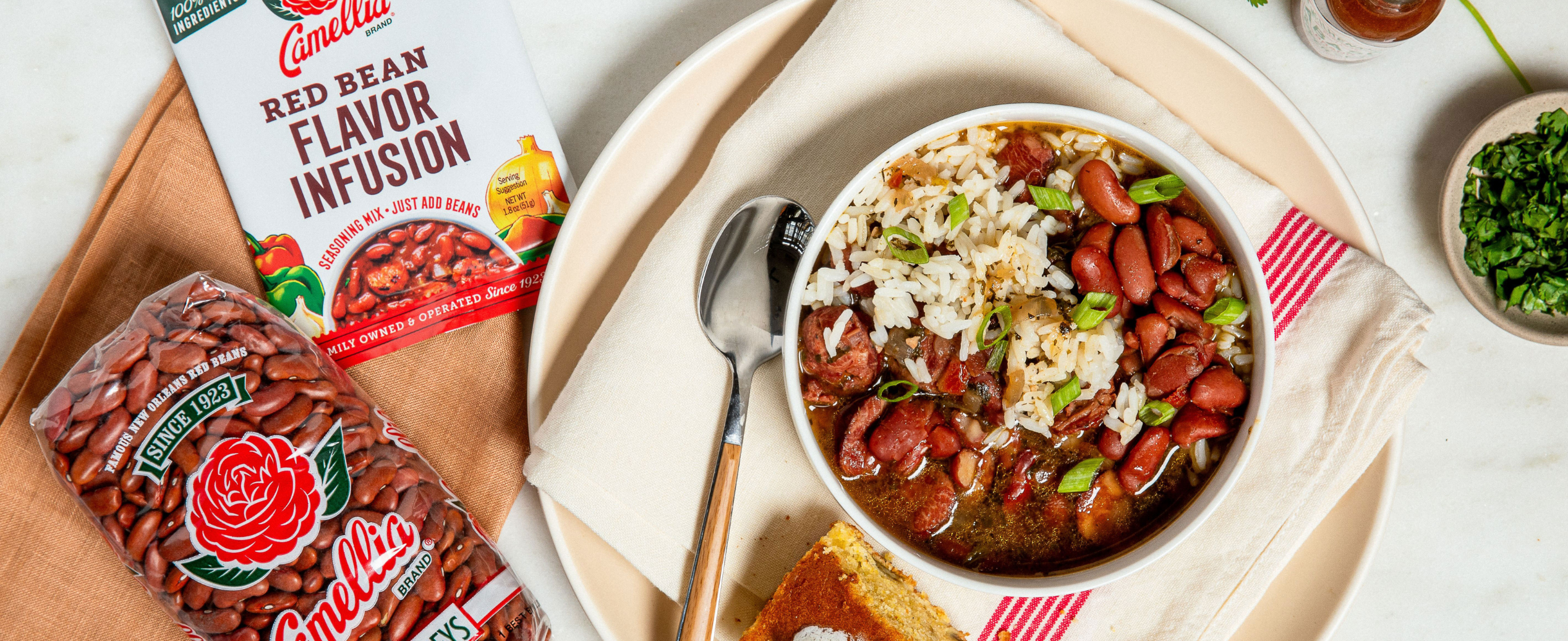 A bowl of red beans and rice garnished with green onions sits on a plate with cornbread. Nearby are Camellia red beans and seasoning mix packages.