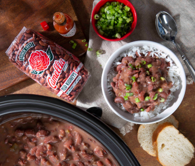 A bowl of red kidney beans over rice garnished with green onions. Nearby are sliced bread, a slow cooker with beans, red pepper sauce, and a bean package.