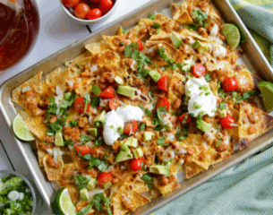 A tray of loaded nachos topped with melted cheese, diced tomatoes, avocado, sour cream, and cilantro. Lime wedges and cherry tomatoes are nearby.