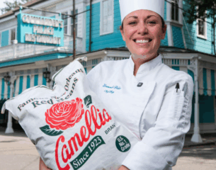 Chef in a white uniform and hat holds a Camellia beans bag, smiling outside Commander's Palace restaurant. The scene conveys pride and tradition.