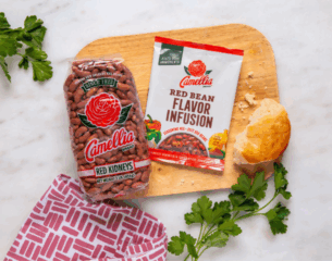 Bag of Camellia red kidney beans and a packet of red bean flavor infusion on a wooden board with a biscuit, parsley, and a patterned cloth nearby.