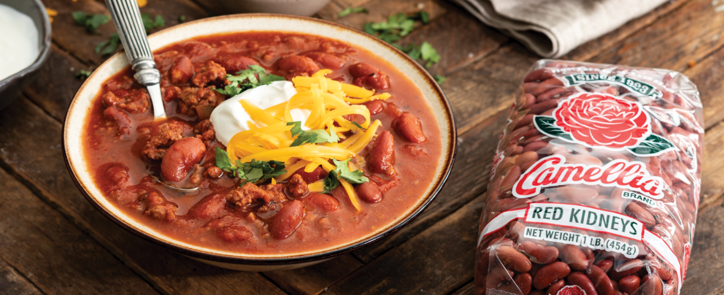 Two bowls of kidney bean chili topped with sour cream, shredded cheese, and cilantro on a wooden table. A bag of Camellia red kidney beans, cornbread, and cheese are nearby, creating a cozy, inviting meal setting.