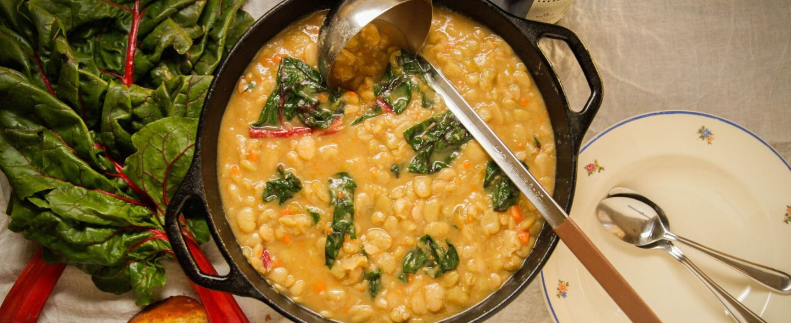 A pot of creamy bean soup with leafy greens sits on a table, accompanied by a serving spoon. Fresh Swiss chard and a plate with utensils are nearby. The scene is warm and inviting.