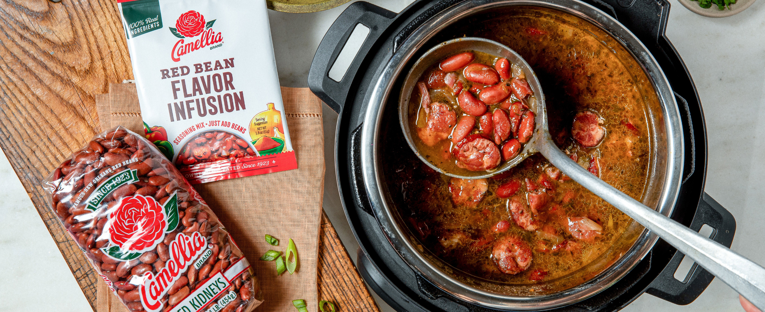 A pot of cooked red beans and sausage stew on a wooden board. Beside it, Camellia red kidney beans and flavor infusion packets are displayed.