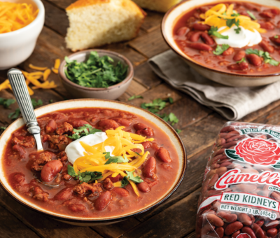 Two bowls of kidney bean chili topped with sour cream, shredded cheese, and cilantro on a wooden table. A bag of Camellia red kidney beans, cornbread, and cheese are nearby, creating a cozy, inviting meal setting.