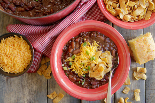 Chili with cheese in a red bowl next to a big pot of chili