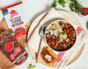 Bowl of red beans and rice garnished with green onions on a plate beside cornbread. A spoon and napkin rest nearby, with packets and cilantro in view.