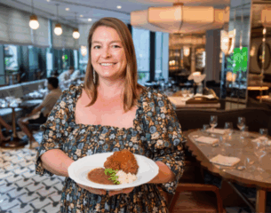 Smiling woman in a floral dress holds a plate with fried chicken, rice, and beans. Elegant restaurant setting with soft lighting and empty tables.