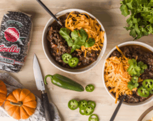 Two bowls of chili topped with cheddar cheese, jalapeño slices, and cilantro are on a wooden table. Nearby are black beans, a knife, jalapeño, mini pumpkins, and cilantro sprigs. The scene feels cozy and inviting.