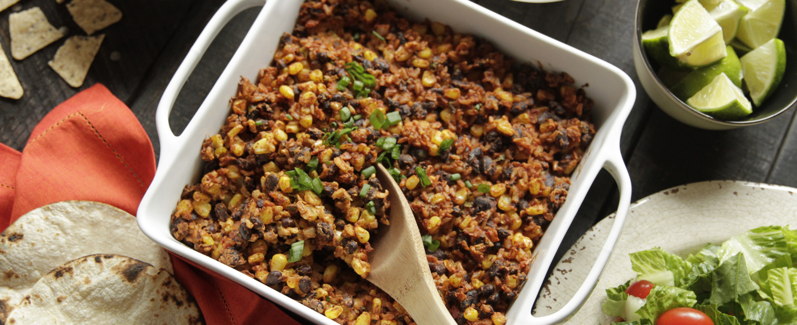 A white dish holds a colorful mix of corn, black beans, and mince with cilantro garnish. Surrounding are tortilla chips, cherry tomatoes, lime wedges, and salad.
