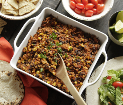 A white dish holds a colorful mix of corn, black beans, and mince with cilantro garnish. Surrounding are tortilla chips, cherry tomatoes, lime wedges, and salad.