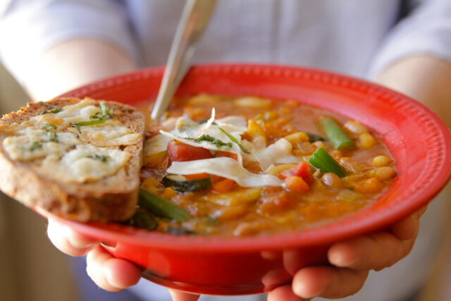 Red bowl of minestrone soup and a piece of bread in someone's hands 