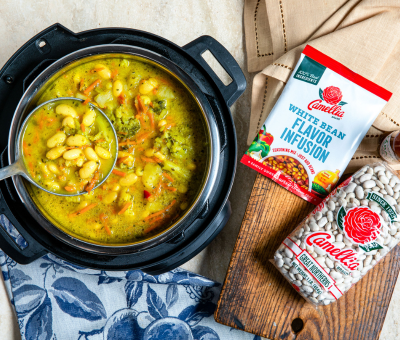 A pot of vibrant bean soup with vegetables, a ladle resting inside. Nearby are packets of white bean flavor infusions and dried beans. Cozy and inviting.