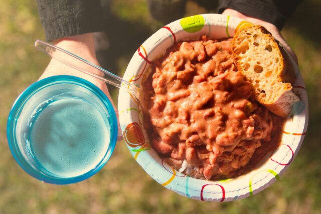 A person holds a colorful paper plate of hearty beans and bread in one hand and a blue cup with a fizzy drink in the other, outdoors on grass.