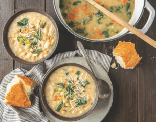 Two bowls of creamy soup with spinach and carrots are garnished with cheese on a wooden table. A pot of soup and rustic bread are nearby, creating a cozy vibe.