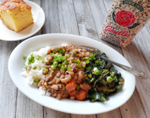A white plate holds a serving of black-eyed peas over rice, garnished with green onions, alongside sautéed greens. A slice of cornbread rests on a small plate next to a bag of Camellia black-eyed peas. The wooden table setting conveys a homely, comforting tone.