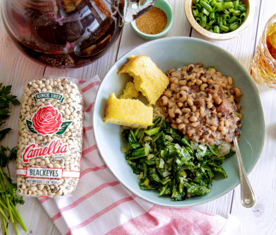 A bowl of black-eyed peas, collard greens, and cornbread on a white table. Nearby, a bag of dry peas, green onions, and tea set a comforting, homey scene.