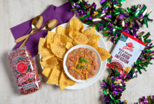 A festive setup with a bowl of bean dip garnished with green onions and cheese, surrounded by tortilla chips, packaging of red kidney beans, flavor infusion mix, and colorful streamers.