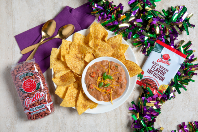 A festive setup with a bowl of bean dip garnished with green onions and cheese, surrounded by tortilla chips, packaging of red kidney beans, flavor infusion mix, and colorful streamers.