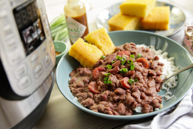 A bowl of red beans and rice topped with green onions and sausage slices, served with cornbread. Nearby are a pressure cooker and hot sauce bottle.