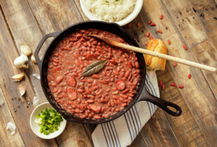 A rustic wooden table with a skillet of vibrant red beans in a rich sauce, topped with a bay leaf and slices of sausage. Nearby, a bowl of rice, chopped green onions, garlic, and crusty bread convey warm, home-cooked comfort.