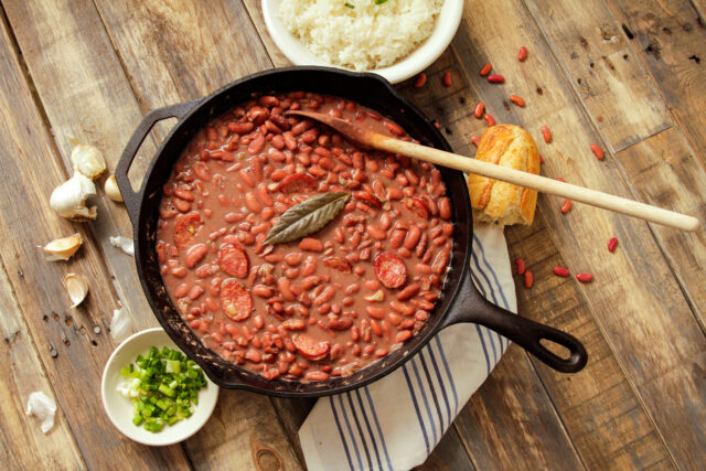 A rustic wooden table with a skillet of vibrant red beans in a rich sauce, topped with a bay leaf and slices of sausage. Nearby, a bowl of rice, chopped green onions, garlic, and crusty bread convey warm, home-cooked comfort.
