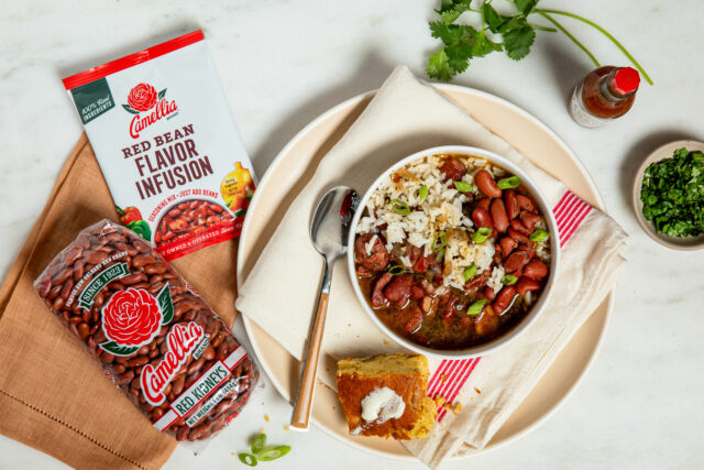 A bowl of rice and red beans with green garnish on a plate, alongside cornbread with butter. Nearby, Camellia red beans packaging, hot sauce, and herbs.