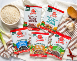 Five Camellia Flavor Infusion bean packets are arranged on a striped cloth. A bowl of rice, garlic, green onion, and a wooden spoon add a rustic touch.