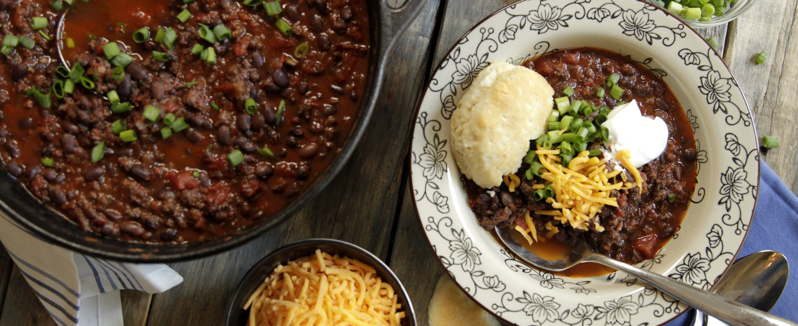 A bowl of chili with beans, cheese, sour cream, and green onions, served with a biscuit. A pot of chili and side of shredded cheese are next to it.
