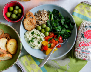 A vibrant bowl with rice, sautéed greens, cherry tomatoes, green olives, and creamy dip, flanked by toasted bread. A sense of fresh, healthy dining.