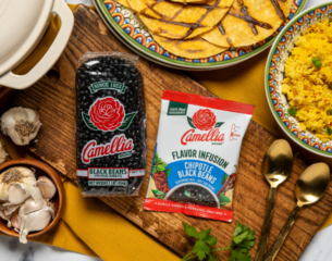A display featuring Camellia Black Beans and Chipotle Black Beans packaging on a wooden board. Nearby are garlic cloves, golden spoons, and colorful plates of sliced tortillas and yellow rice, creating a vibrant, appetizing scene.